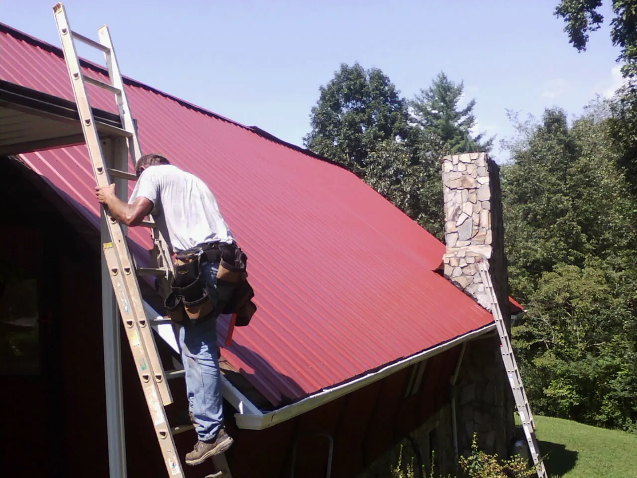 Roofer on a ladder beside a red metal roof and stone chimney — Soffit & Fascia Repair by Roofing Brothers in Newington